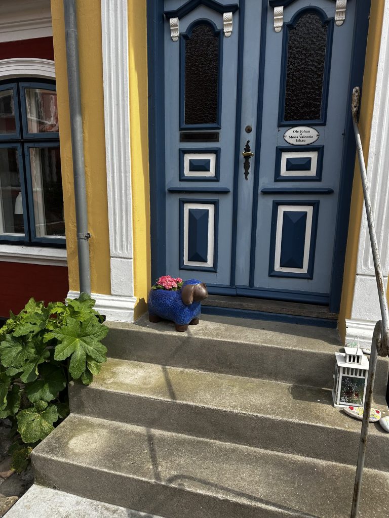 Blaue Doppeltür mit Messingklopfer und Namensschild, davor eine Tonfigur mit Blumentopf